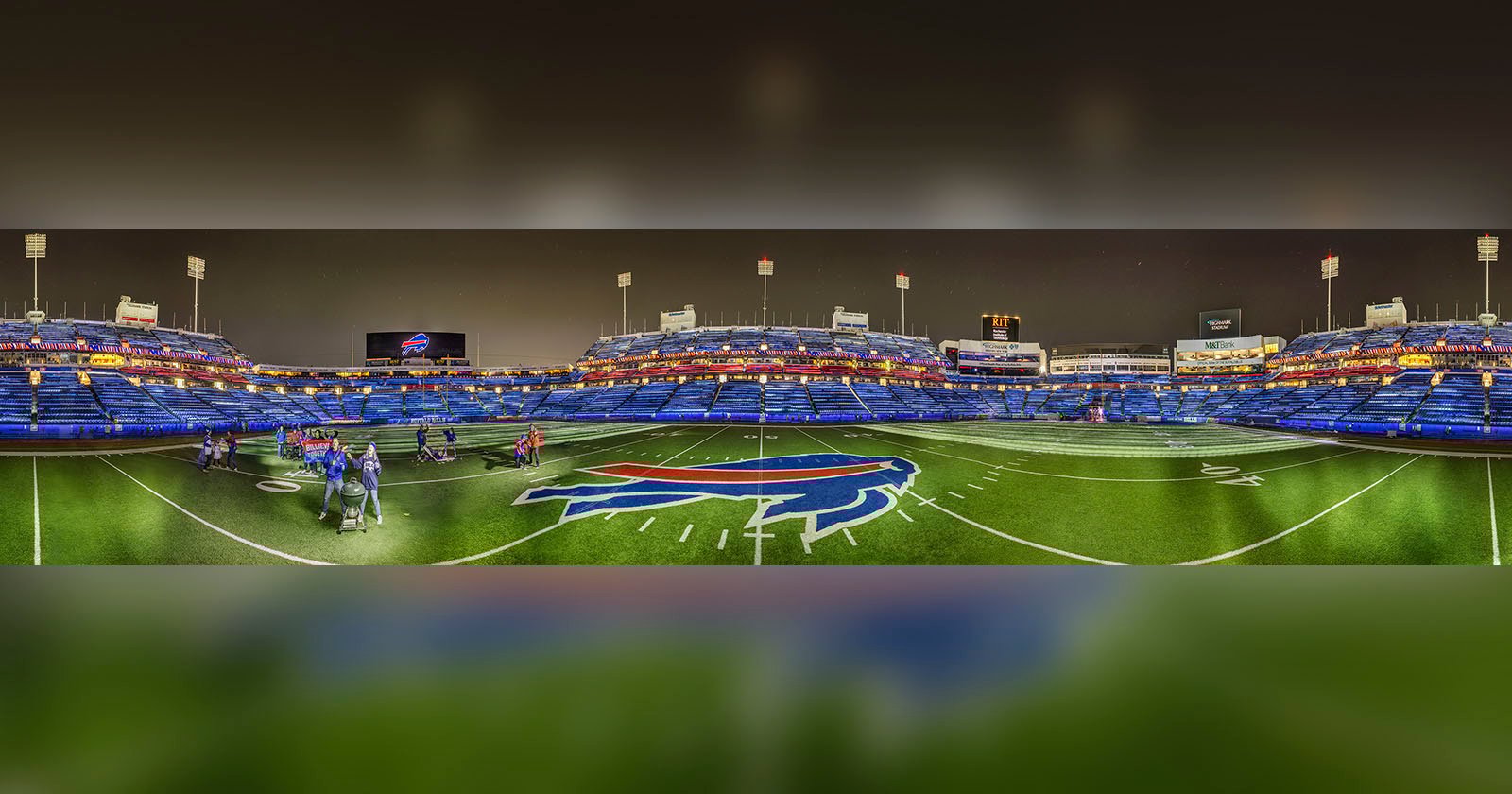 A wide-angle view of an empty football stadium at night, with bright lights and a large buffalo logo at midfield. A few people stand on the turf near the center. Blue seats fill the stands in the background. A wide-angle view of an empty football stadium at night, with bright lights and a large buffalo logo at midfield. A few people stand on the turf near the center. Blue seats fill the stands in the background.