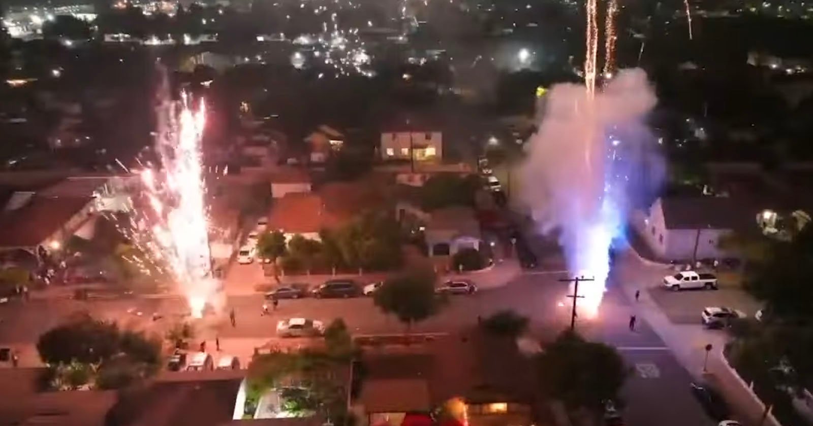 Aerial view of a residential neighborhood at night with fireworks shooting up from the street, illuminating houses and trees while city lights glow in the background. Aerial view of a residential neighborhood at night with fireworks shooting up from the street, illuminating houses and trees while city lights glow in the background.
