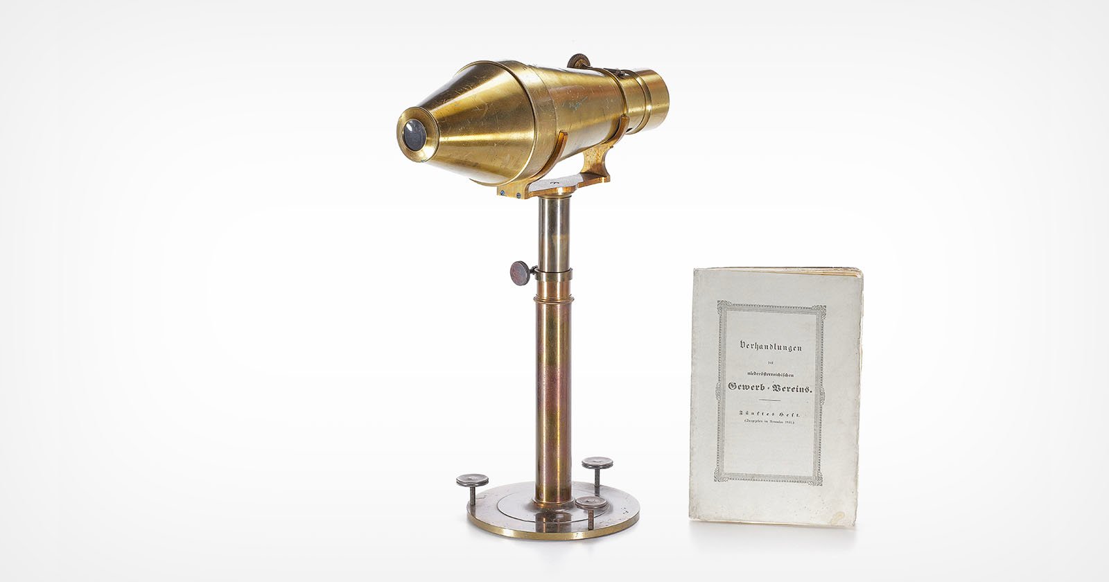 A vintage brass microscope on a metal stand beside an old booklet with a gray border and German text on the cover, both placed against a white background. A vintage brass microscope on a metal stand beside an old booklet with a gray border and German text on the cover, both placed against a white background.