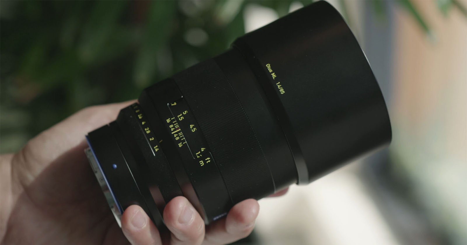 A hand holding a black camera lens with yellow markings and numbers, against a blurred background with green leaves and natural light. A hand holding a black camera lens with yellow markings and numbers, against a blurred background with green leaves and natural light.